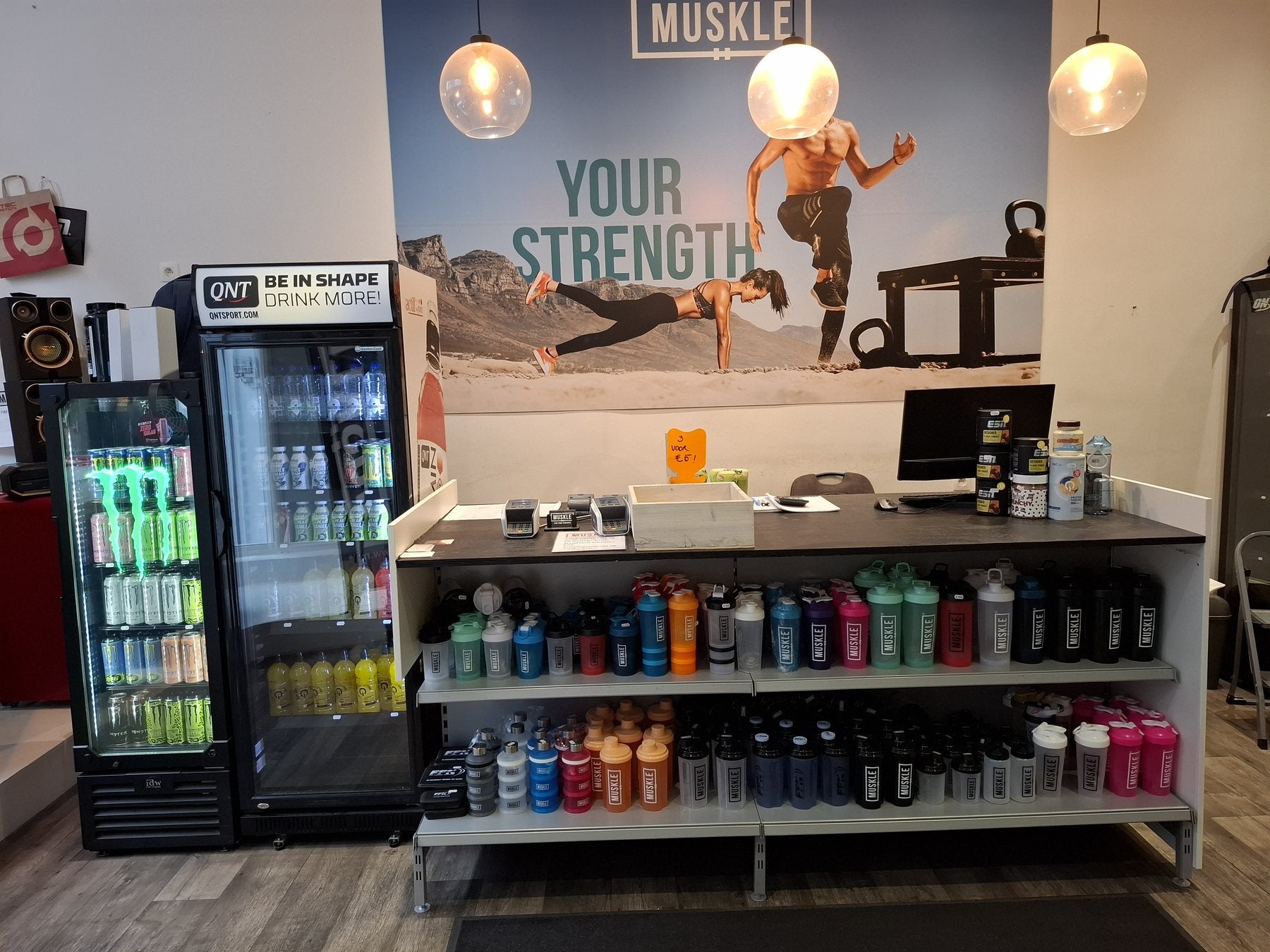 Servicebalie van MUSKLE Antwerpen met shakers, frisdrankkoelkasten en YOUR STRENGTH-muur